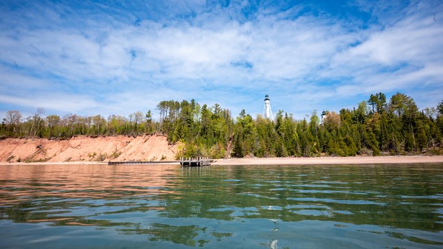 Photograph of sunny day on lake with lighthouse, forest, and sandstone cliffs in background.