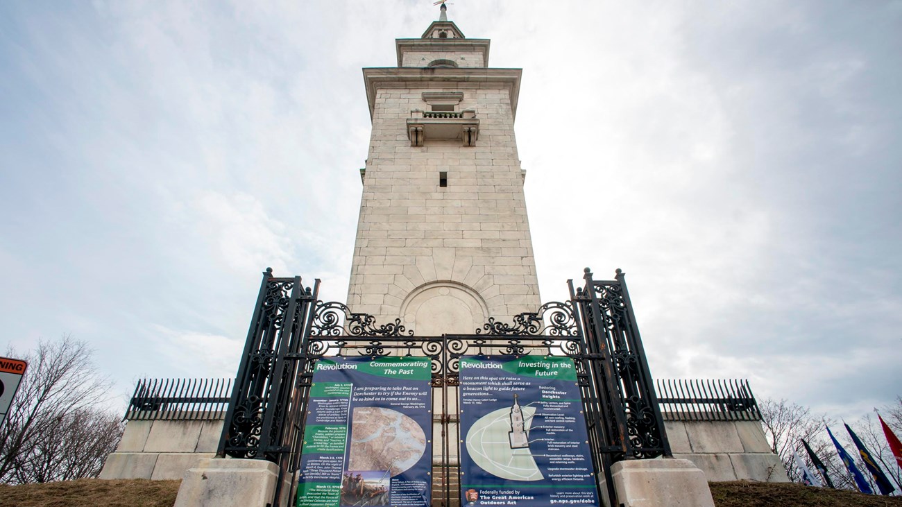 Dorchester Heights Monument with stage and podium in front and audience sitting on sidewalk sides.