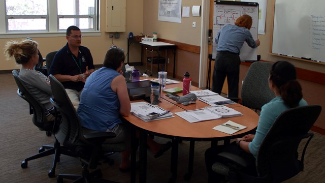 Several people seated at an oval table in front of a white board and windows in back