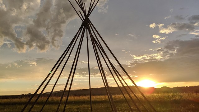 several long poles arranged in a tipi shape against a sunset and badlands buttes in background.