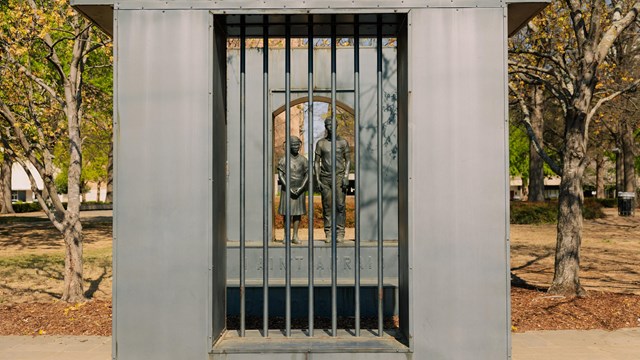 A metal statue of two children seen through iron bars. A path runs between the children and bars.
