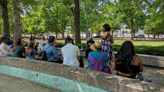 A speaker speaks into a microphone. A group of 14 people sit on stone benches listening.