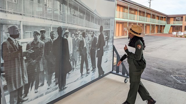 A ranger stands looks at an 8 foot tall rolling mural. A motel is seen above and in the background. 