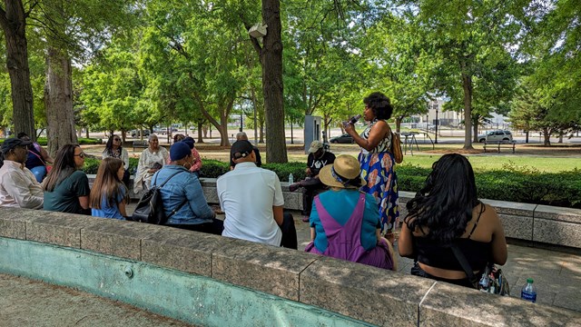 A person stands in a park holding a microphone. A group sits on stone benches listening.