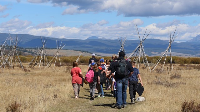 A group of kids and adults hike across native grass toward tipis and the mountains beyond. 