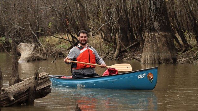 A man wearing an orange life vest paddling a blue canoe in murky water past leafless trees.