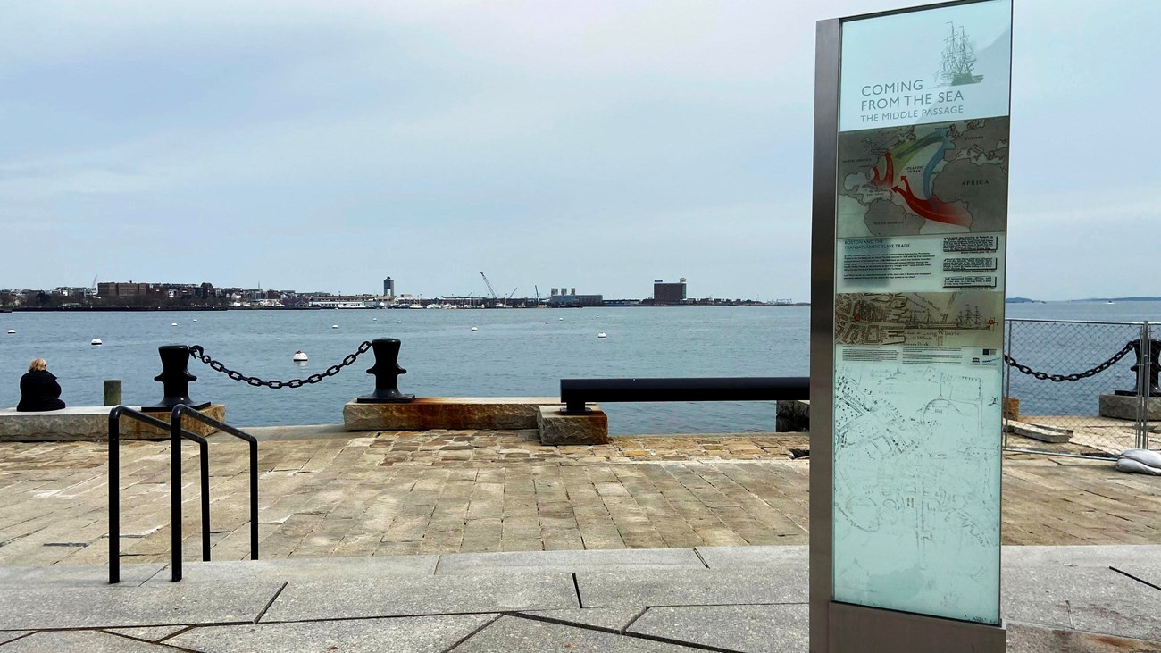 View of the end of a wharf with harbor in the background; on the right is a rectangular wayside.