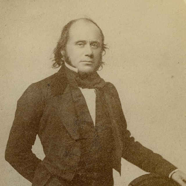 Man poses with his hand resting on the top of a chair. He wears a dress coat with a high collar.