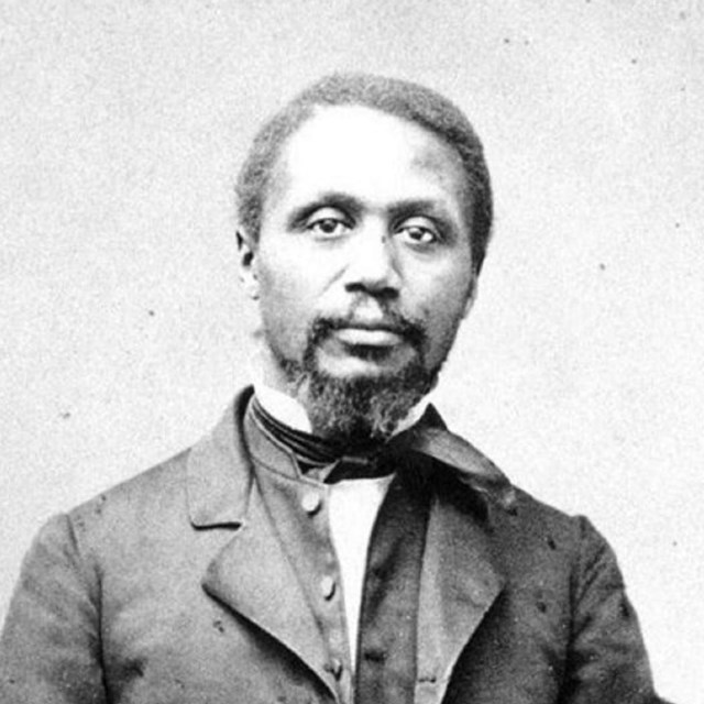 African American man standing next to a column, resting his left arm on the the column. 