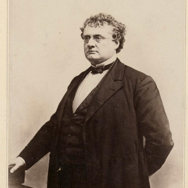 White man with short curly hair & glasses, wearing a dark three-piece suit standing next to a desk. 