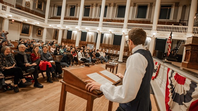 A Ranger dressed in colonial kit stands at a podium and speaks to people seated in a great hall.