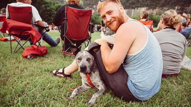 A man sits with his dog. Groups of people sit behind them.