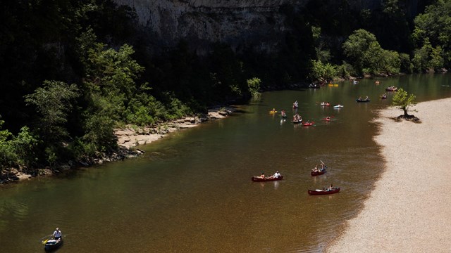 A river flows by green bluffs with people in kayaks floating by and stopped on a gravel bar..