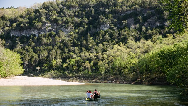 A canoe with two paddlers flows down Buffalo River. They paddle towards a tree-covered bluff line.