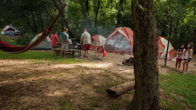 A group of campers gathers by a picnic table. Two other campers stand near a fire pit and tents.