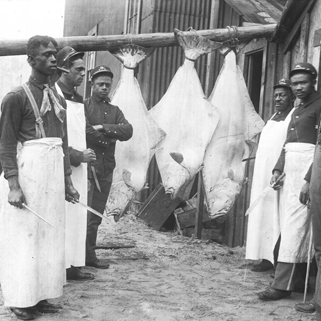 Six soldiers stand and pose for a photo with three giant fish in the background