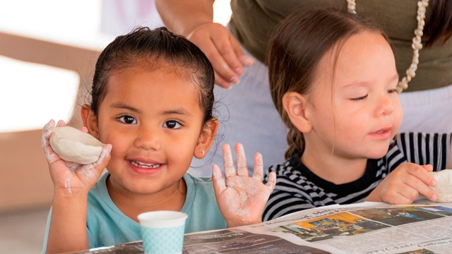 A student holds up a clay pinch pot while another student works on a piece of clay 