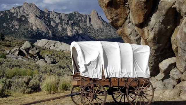 A covered wagon sits in front of a large rock outcropping.