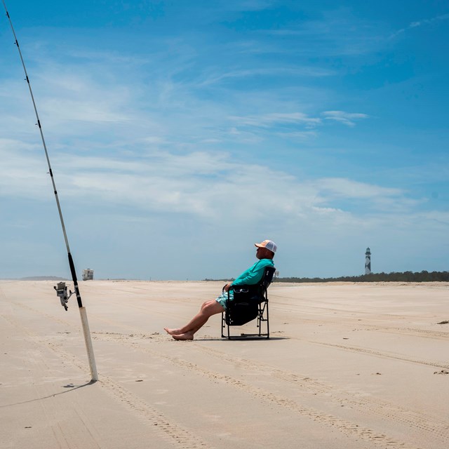A angler sits on a chair with fishing poles in front of him. 