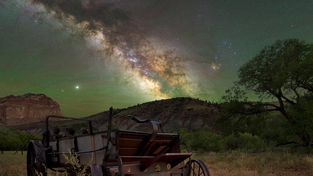 The Milky Way over an old wagon with cliffs and trees in the background