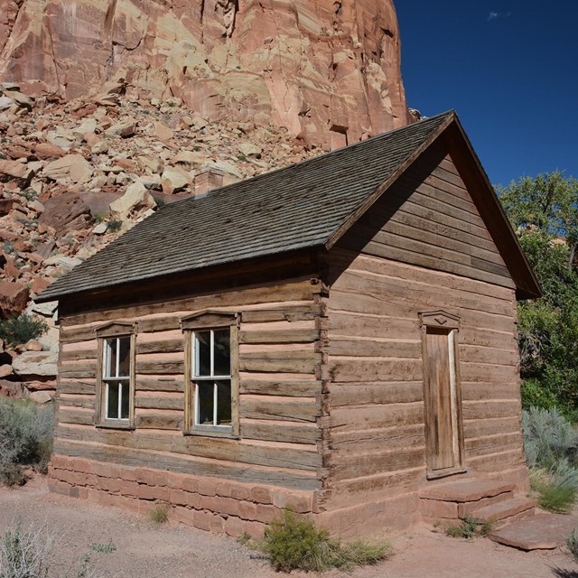 Small log cabin-like building with windows and a door, below red rock cliffs and blue sky.