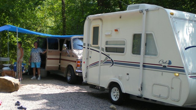 People stand under a tarp canopy near a van and a small camper