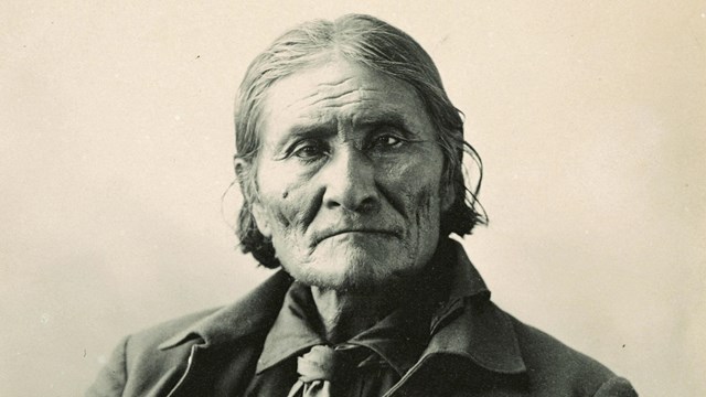 Sepia-toned photograph of seated man wearing a jacket, with hair cropped above his shoulder. 