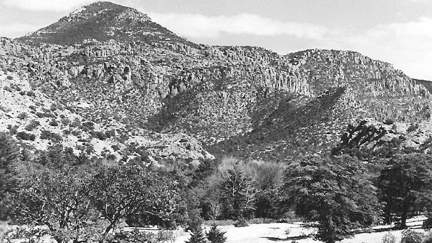 Black and white photograph of field and trees in front of tall hills with rock pinnacles. 