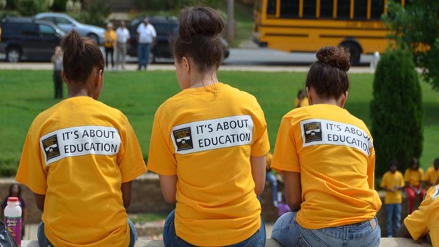 Three students in yellow shirts reading “It’s About Education” sit together near a school bus.