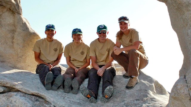 Four crew members pose sitting under a granite arch.