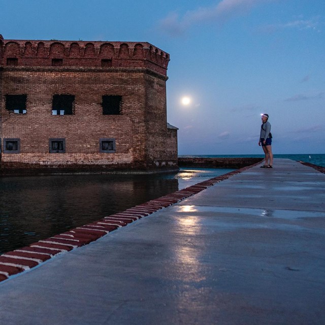 A person with a headlamp gazes at an old brick fort at night from a platform surrounded by water