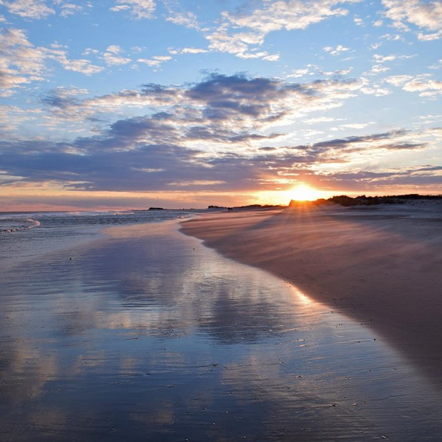 Sun sets over a brown sandy beach with orange and purple clouds dotting the sky