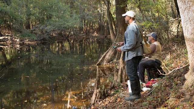 Two people fishing, one standing, one sitting, along water's edge