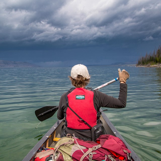 a boy in a red lifejacket paddles a canoe on a lake