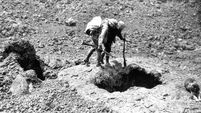 black and white photo of two men with packs and rifles looking into a hole while a dog looks on