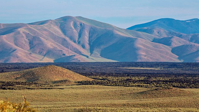 an open area covered in sagebrush surrounded by black lava rocks and mountains in the distance