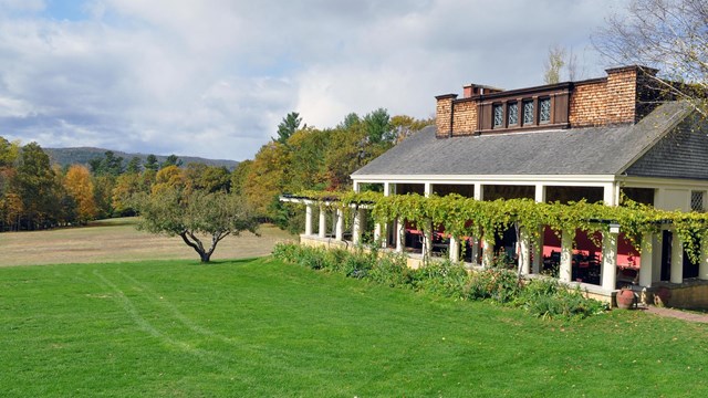 Vines crawl over a trellis that surrounds a long, one-story structure in a rural landscape.