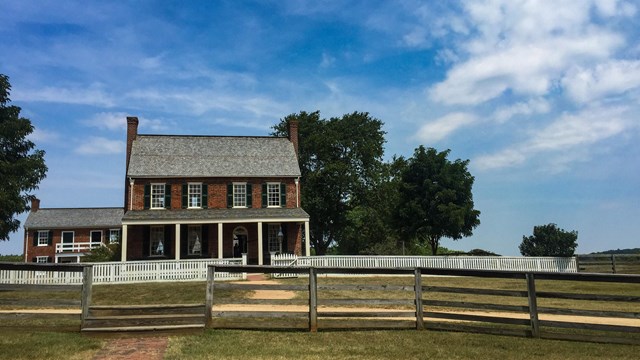 A two-story red brick house behind a picket fence and a wooden fence