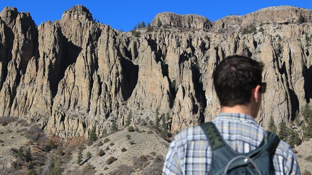 Person wearing a backpack and flannel shirt standing in front of tall pinnacle spires
