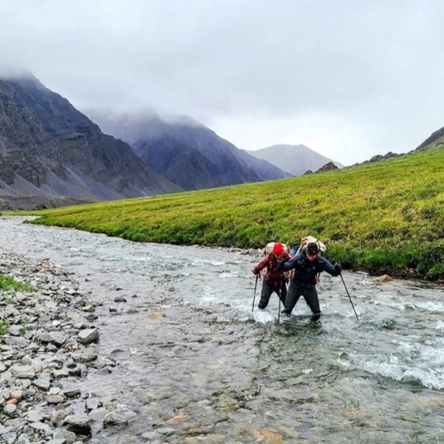 Two people wearing backpacks ford a river at the base of large, rocky mountains.