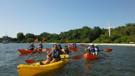A park ranger on kayak leads others on the water. A large cross is in the background.