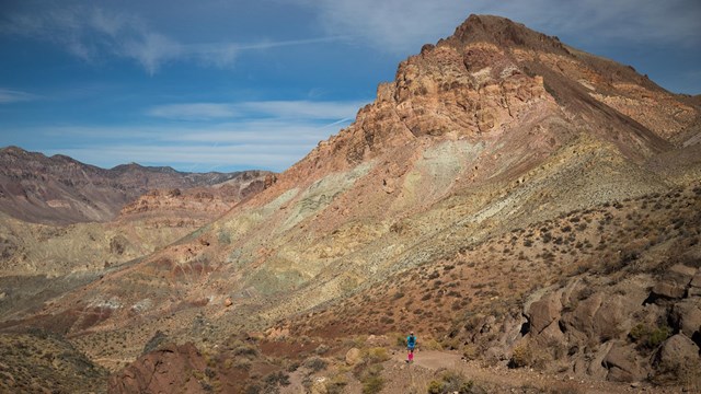 A runner on a dirt road, set in a dramatic desert setting, with multi-hued rocks, distant mountains.