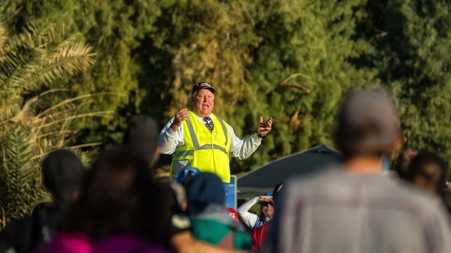 A man in a bright jacket stands above a crowd of people and speaks to them