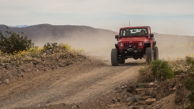 A bright red Jeep, slightly raised on its suspension, drives toward the camera down a dusty road.