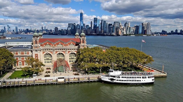 An aerial view of the main immigration building at Ellis Island, the NYC skyline is behind