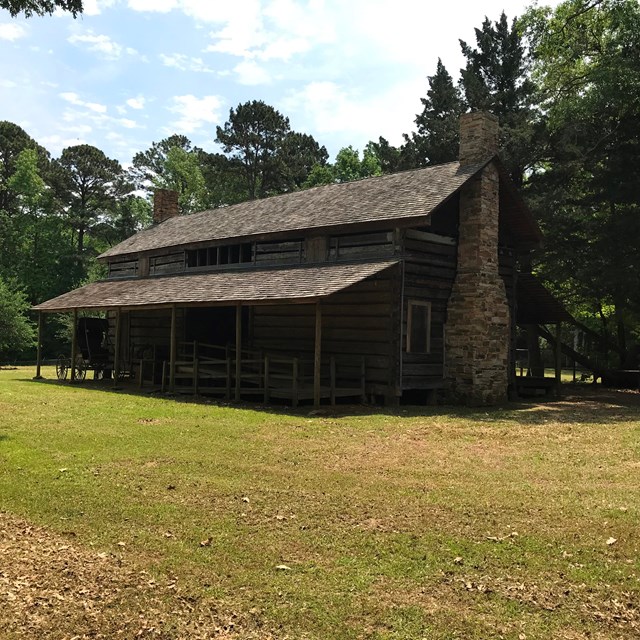 A wooden long house set on a grassy field, surrounded by trees.