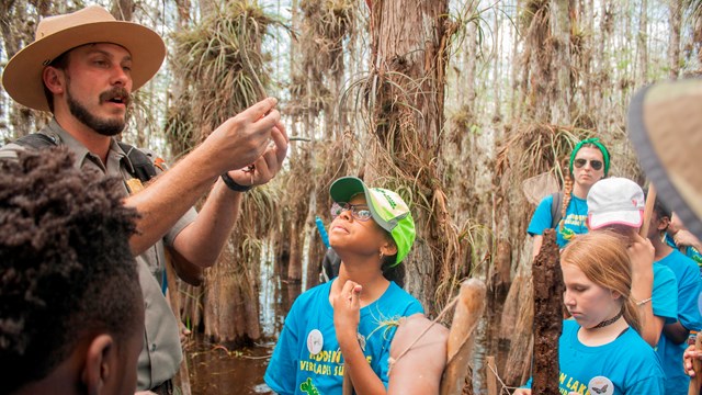 A ranger holds plants up, while students look, while standing in water and surrounded by trees