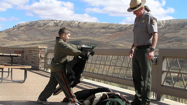 A child places a coat on top of a pile of coats while a ranger watches.