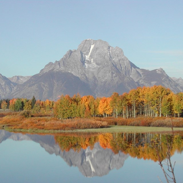 Mountains with some snow and aspens changing color reflected in water below.