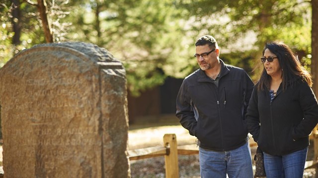Two visitors look at a vertical stone monument with green forest areas behind them